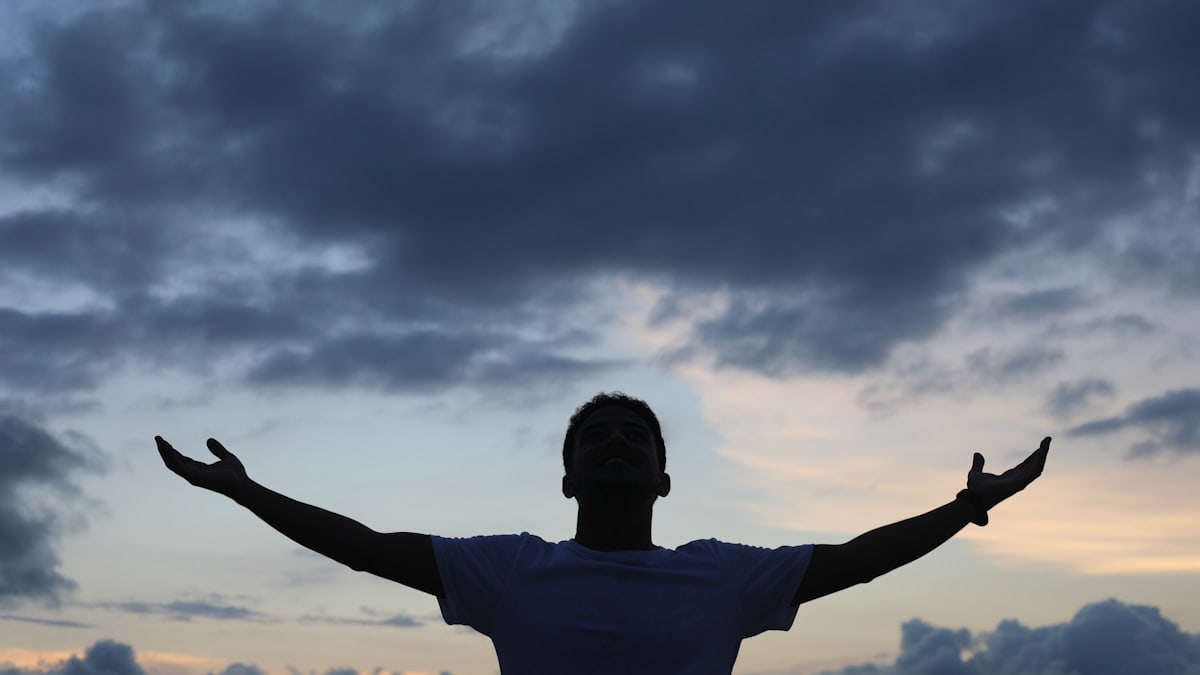 man in white t-shirt raising his hands under cloudy sky during daytime